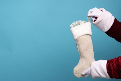 Photo of Santa Claus holding stocking with dollar banknotes on light blue background, closeup. Space for text