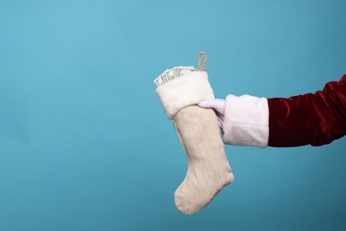 Photo of Santa Claus holding stocking with dollar banknotes on light blue background, closeup. Space for text