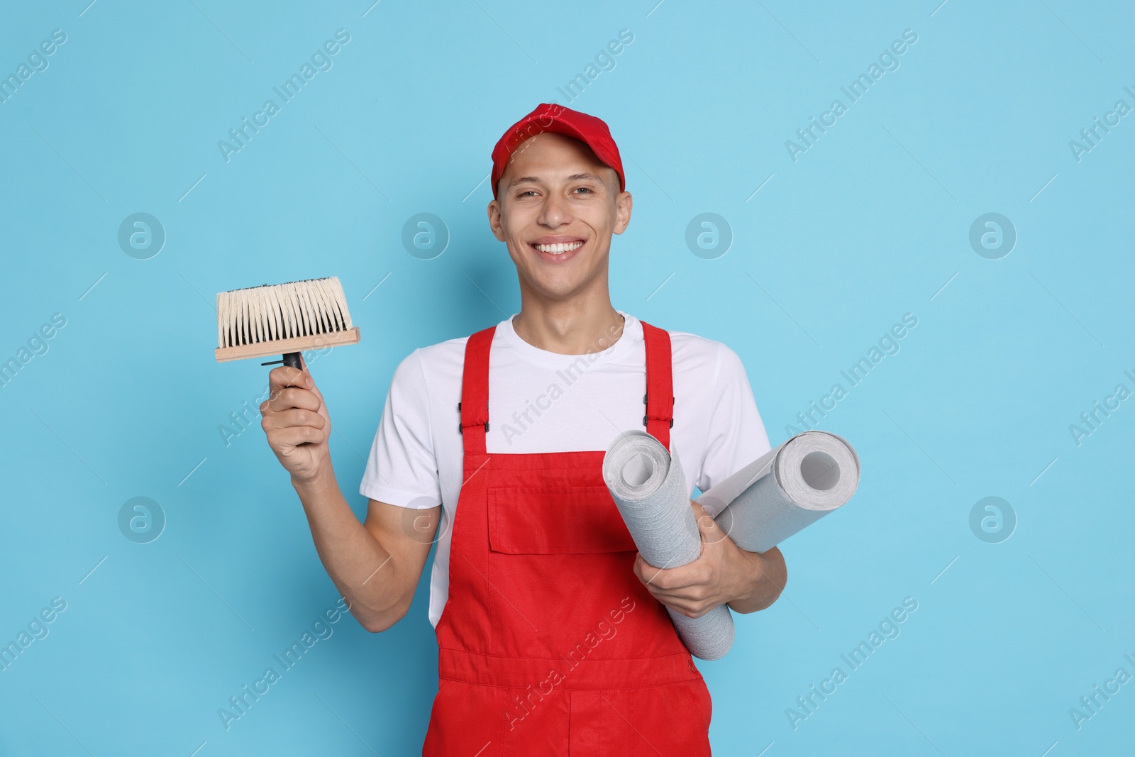 Photo of Young decorator with brush and rolls of wallpaper on light blue background