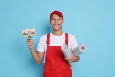 Young decorator with brush and rolls of wallpaper on light blue background