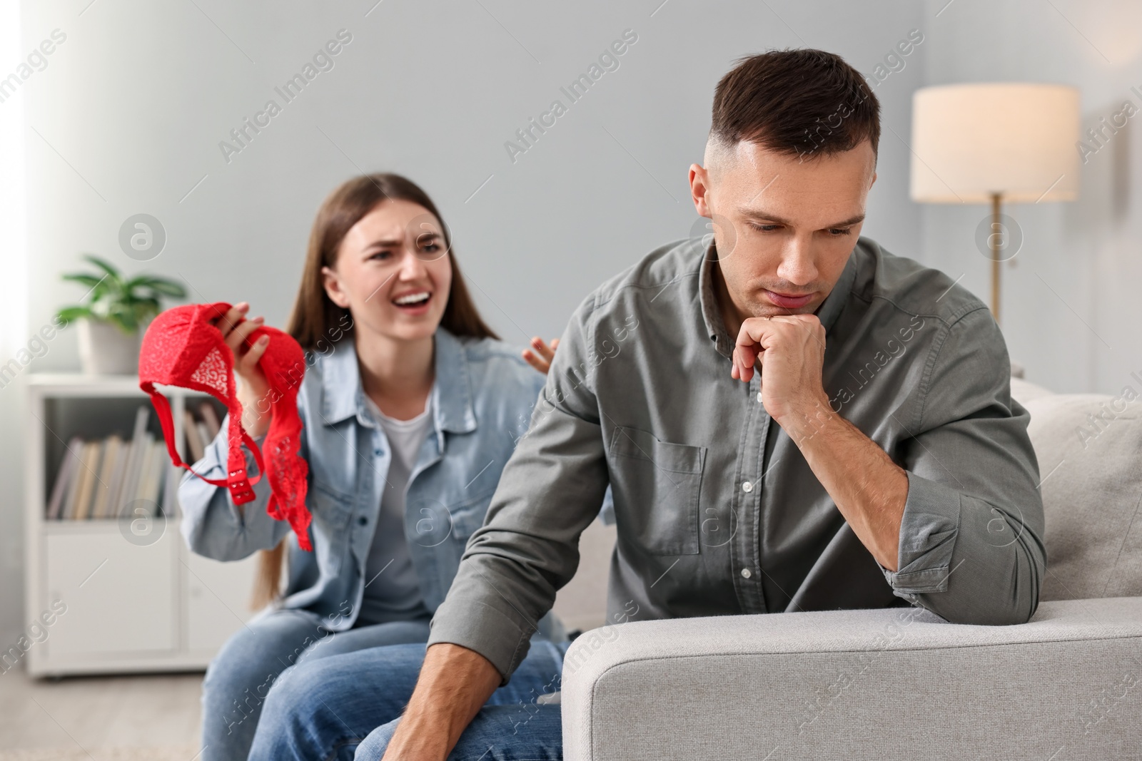 Photo of Emotional wife with mistress bra as proof screaming at her unfaithful husband while he ignoring her indoors, selective focus