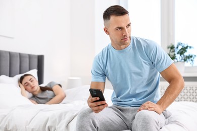 Jealous man checking wife's smartphone while she sleeping in bedroom, selective focus