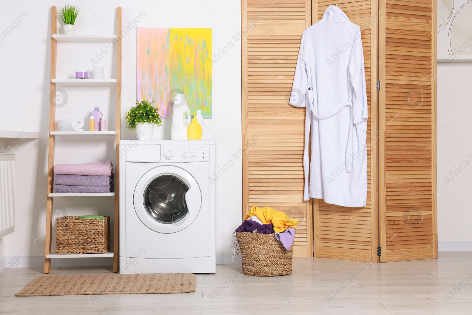 Photo of Washing machine, detergents, bathrobe, clean towels and basket with laundry in room