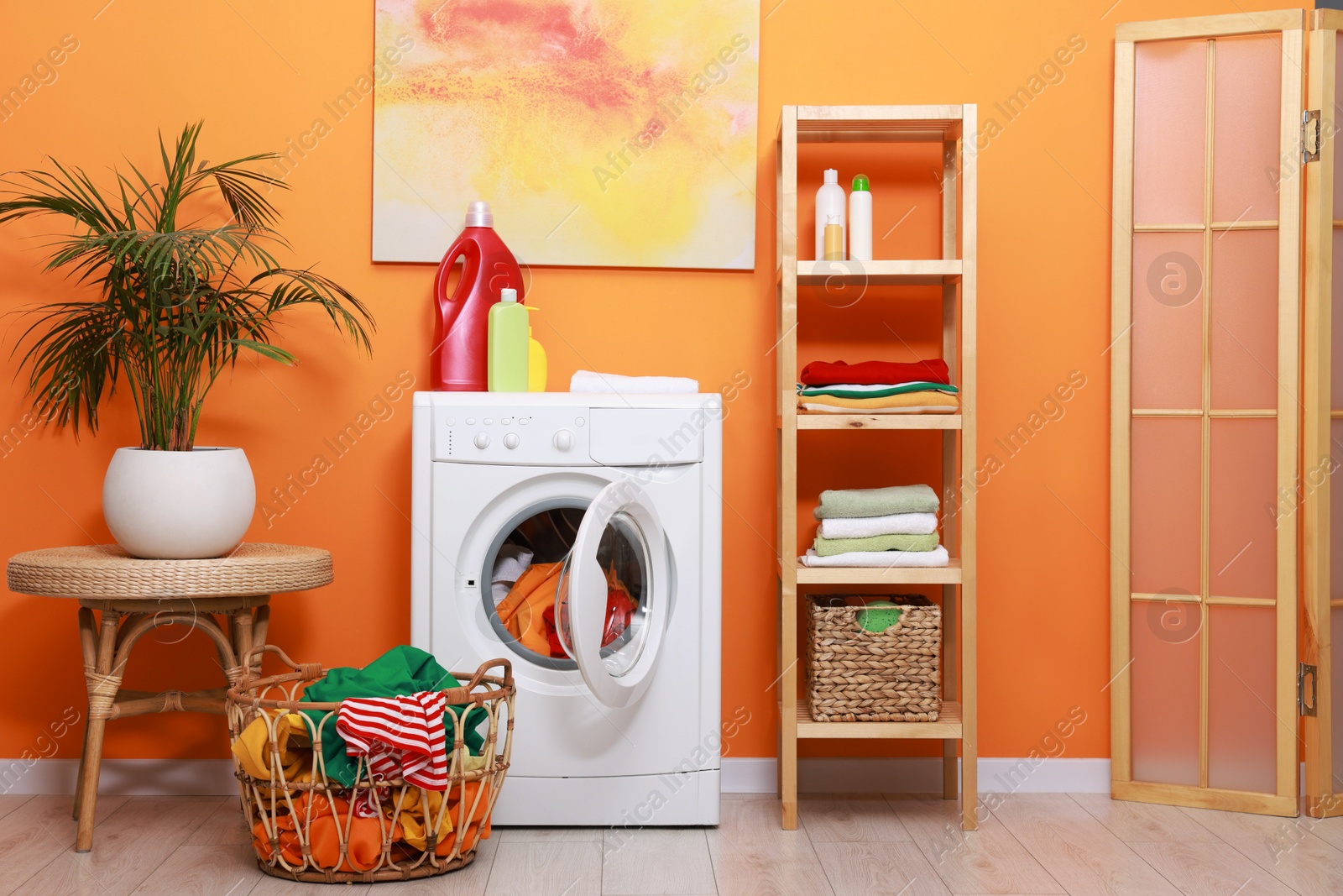 Photo of Washing machine, detergents, clean towels and basket with laundry near orange wall indoors