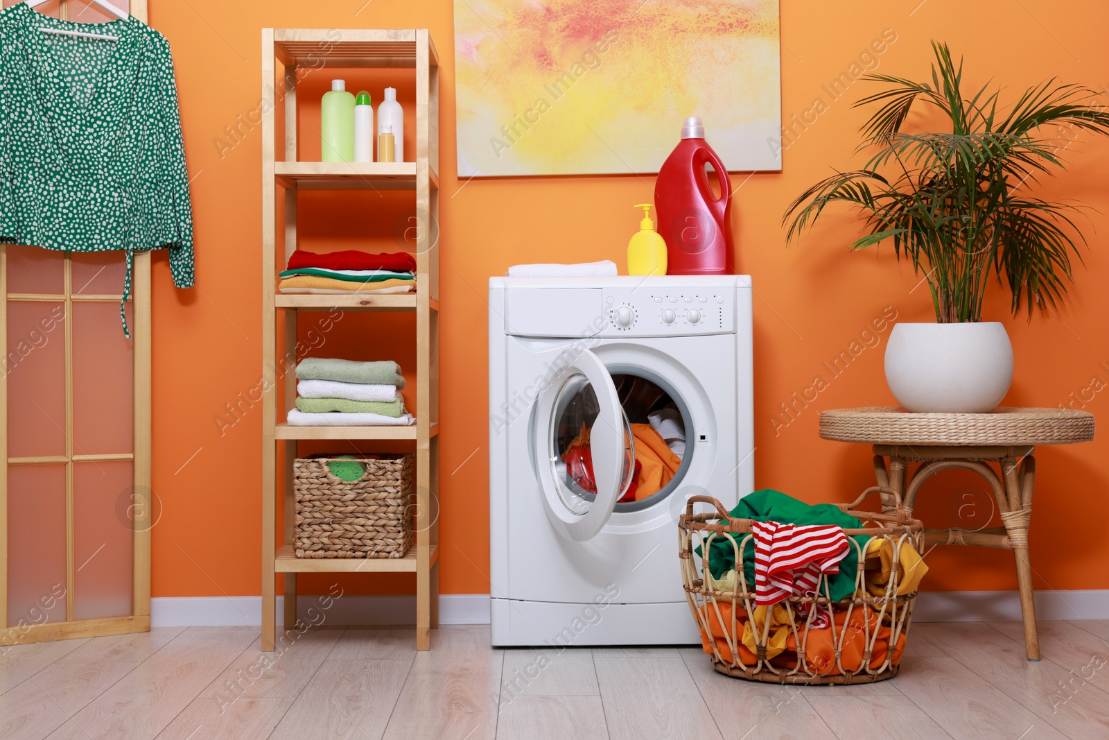 Photo of Washing machine, detergents, clean towels and basket with laundry near orange wall indoors