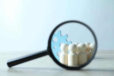 Photo of Human resources concept. Looking at wooden figures and gearwheel through magnifying glass on table against light background, closeup. Space for text