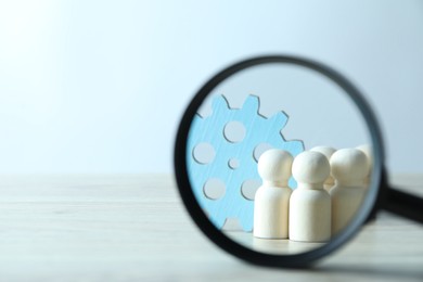 Photo of Human resources concept. Looking at wooden figures and gearwheel through magnifying glass on table against light background, closeup. Space for text