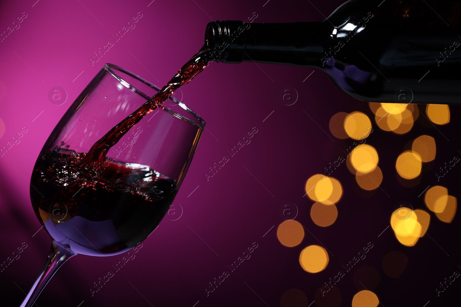 Photo of Pouring red wine into glass against purple background, closeup. Bokeh effect