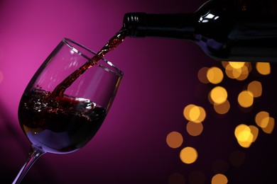 Photo of Pouring red wine into glass against purple background, closeup. Bokeh effect