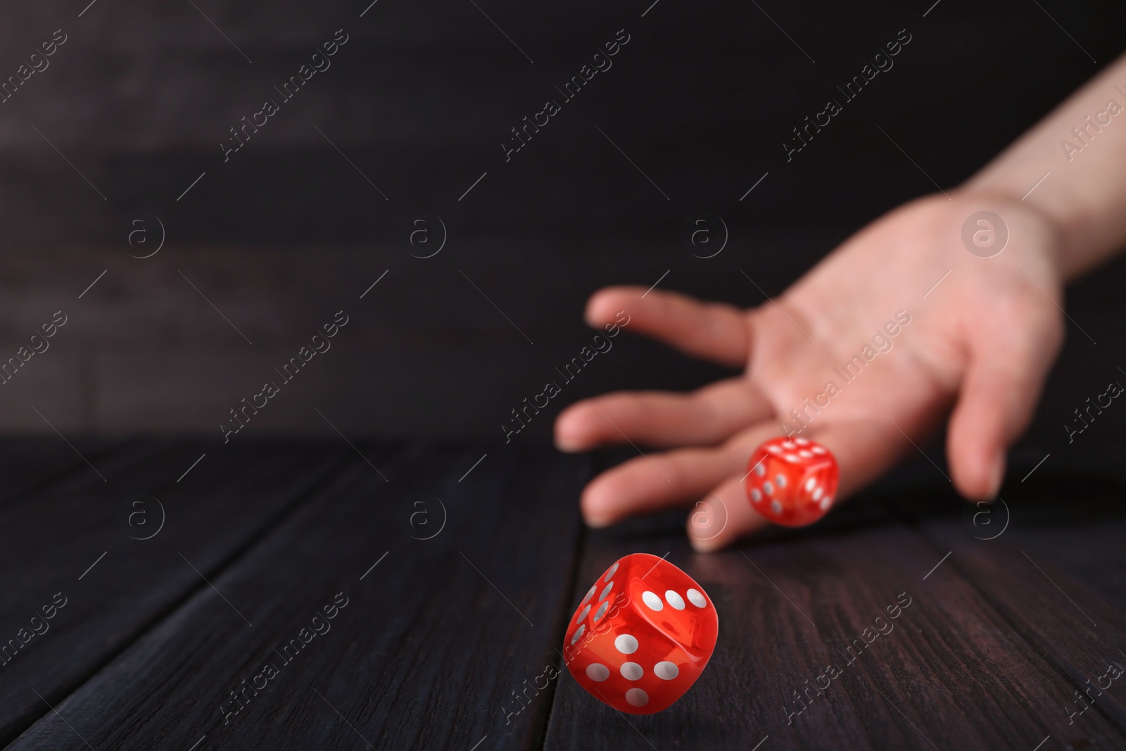 Image of Woman throwing red dice on black wooden table, closeup. Space for text