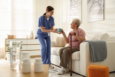 Photo of Caregiver giving cup of drink to senior woman indoors. Home health care service