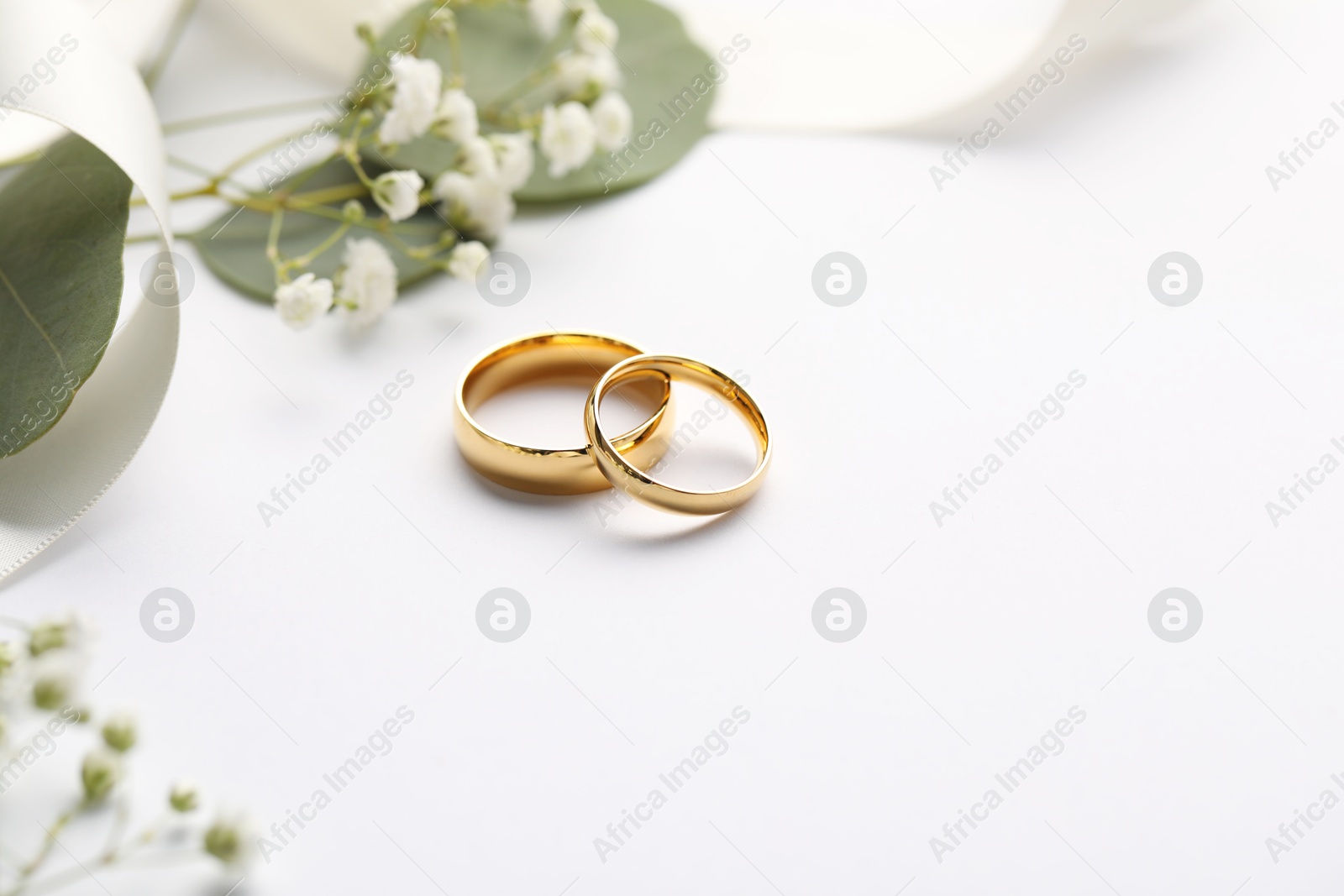 Photo of Golden wedding rings, ribbon, leaves and flowers on white background, closeup