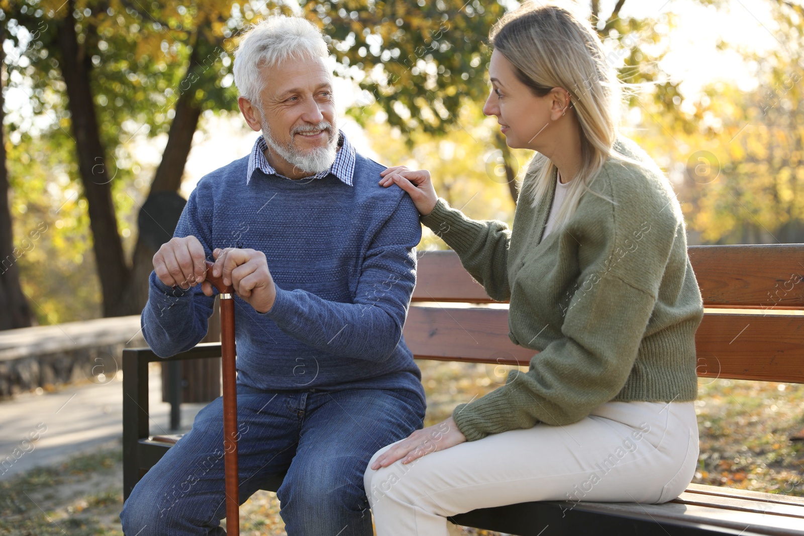 Photo of Caregiver assisting senior man on wooden bench in park. Home health care service