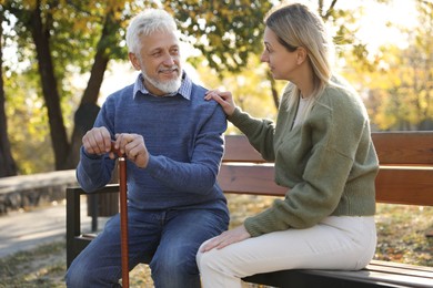 Photo of Caregiver assisting senior man on wooden bench in park. Home health care service