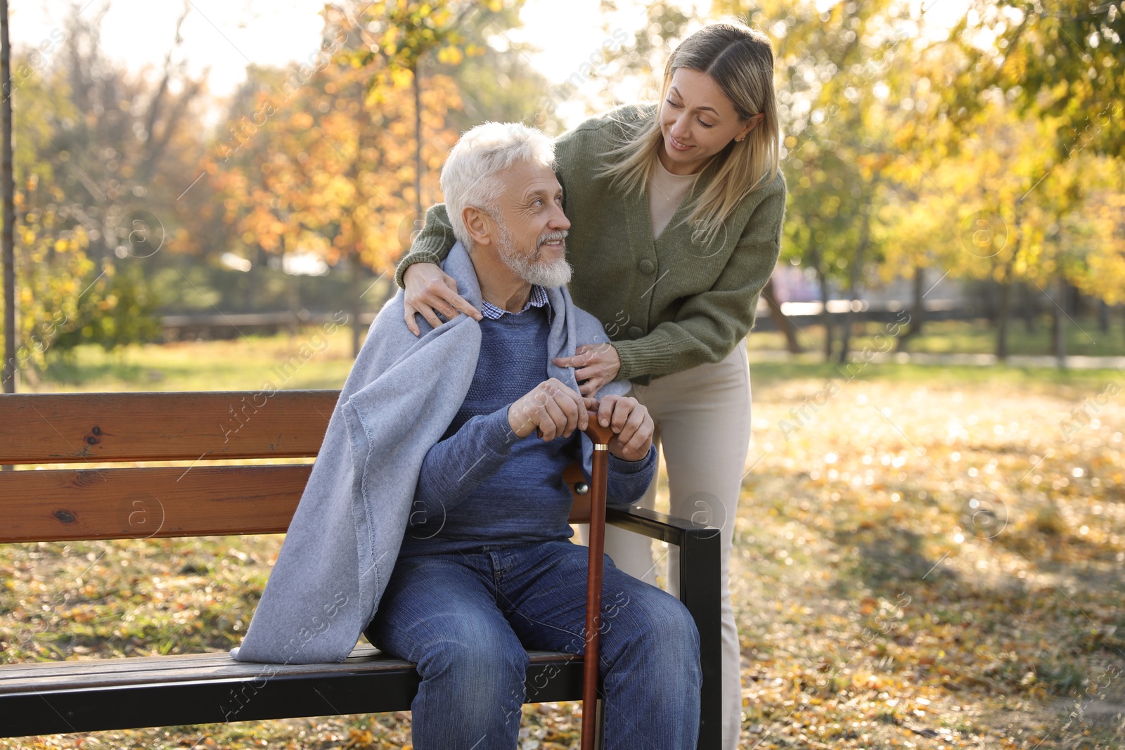 Photo of Caregiver assisting senior man on wooden bench in park. Home health care service