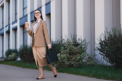 Photo of Smiling businesswoman in stylish suit with paper cup walking outdoors. Space for text