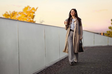 Smiling businesswoman in stylish suit with paper cup walking outdoors. Space for text