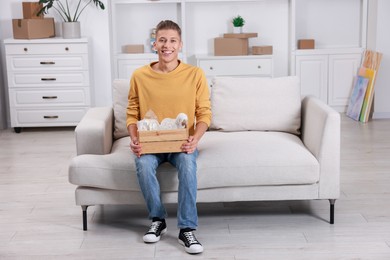 Photo of Happy man holding wooden crate with stuff on sofa in new apartment. Housewarming party