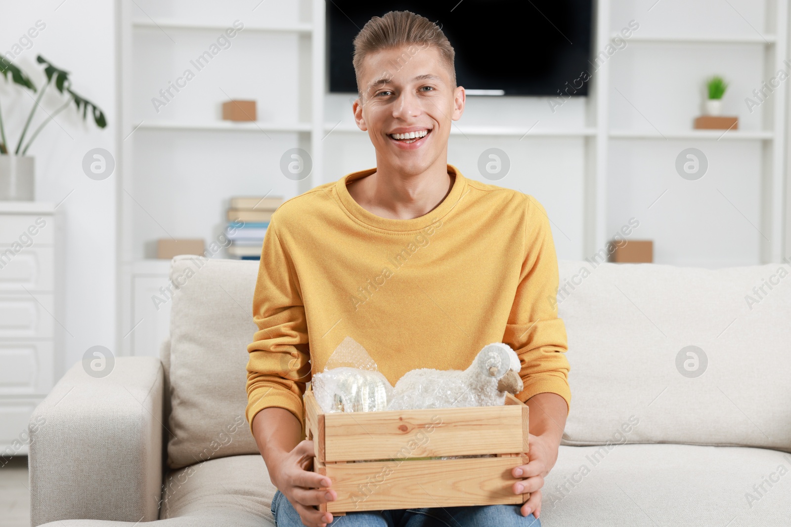 Photo of Happy man holding wooden crate with stuff on sofa in new apartment. Housewarming party