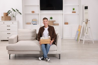 Photo of Happy man holding wooden crate with stuff on sofa in new apartment. Housewarming party