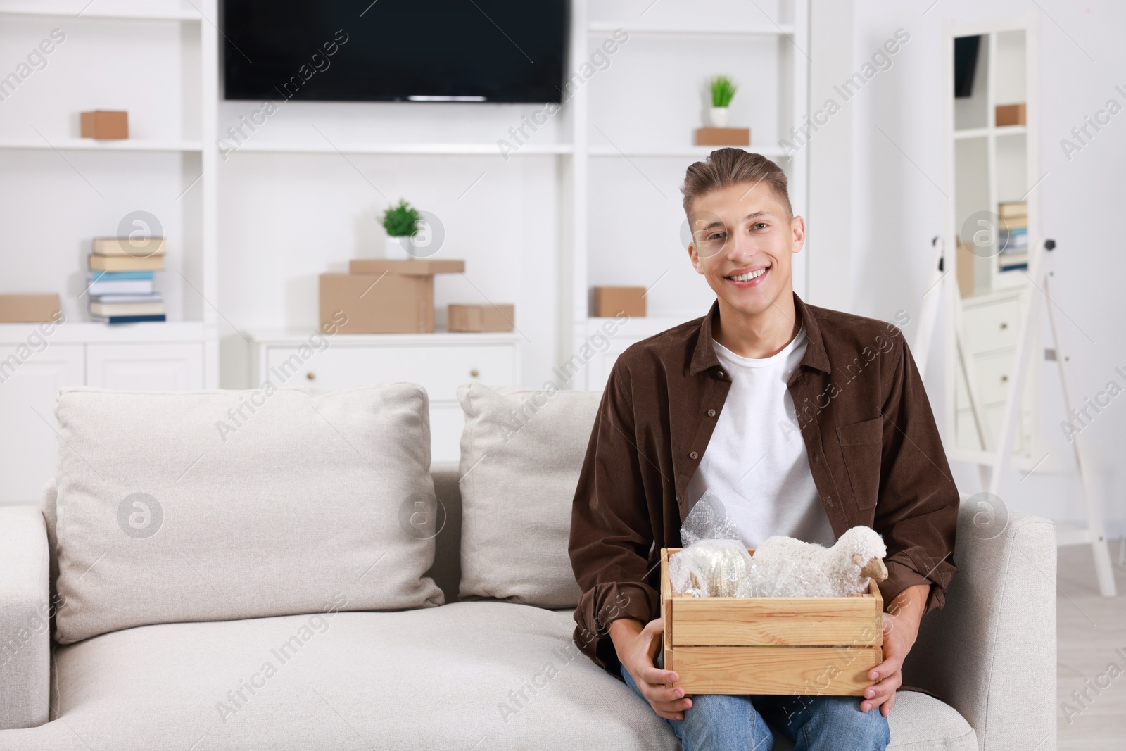 Photo of Happy man holding wooden crate with stuff on sofa in new apartment. Housewarming party