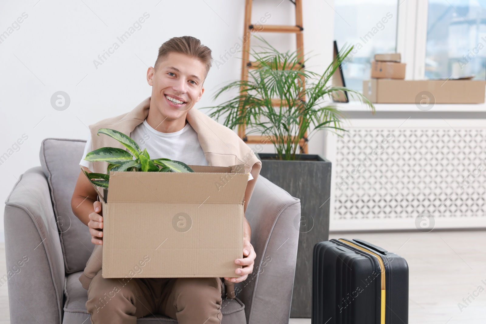 Photo of Happy man holding moving box with houseplant in new apartment. Housewarming party