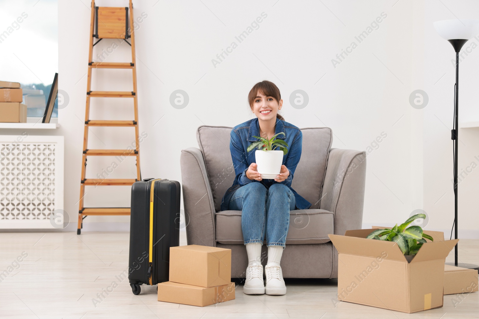 Photo of Happy woman with houseplant, moving boxes and suitcase in new apartment. Housewarming party