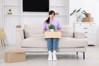 Photo of Happy woman holding wooden crate with stuff on sofa in new apartment. Housewarming party