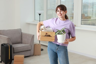 Photo of Happy woman holding wooden crate with stuff and houseplant in new apartment. Housewarming party