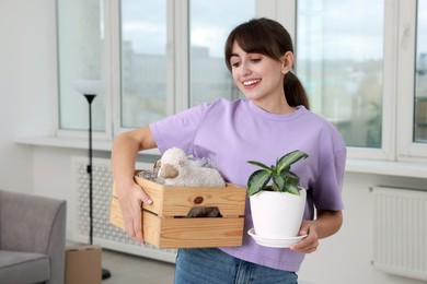 Happy woman holding wooden crate with stuff and houseplant in new apartment. Housewarming party