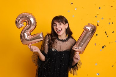 Photo of Coming of age party - 21st birthday. Young woman holding number shaped balloons on yellow background with confetti