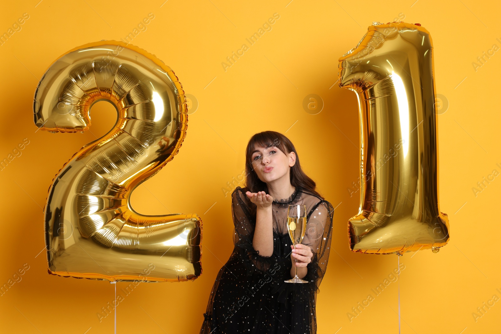 Photo of Coming of age party - 21st birthday. Young woman with number shaped balloons and glass of wine blowing kiss on yellow background