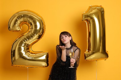 Coming of age party - 21st birthday. Young woman with number shaped balloons and glass of wine blowing kiss on yellow background