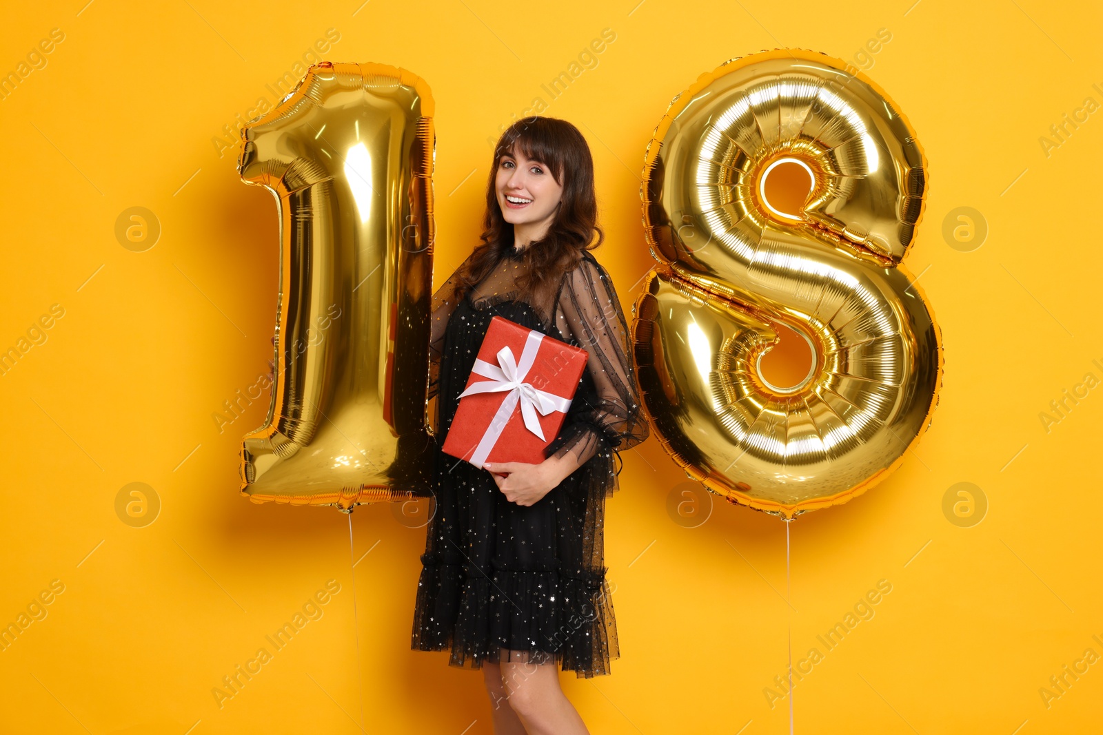 Photo of Coming of age party - 18th birthday. Young woman with number shaped balloons and gift box on yellow background