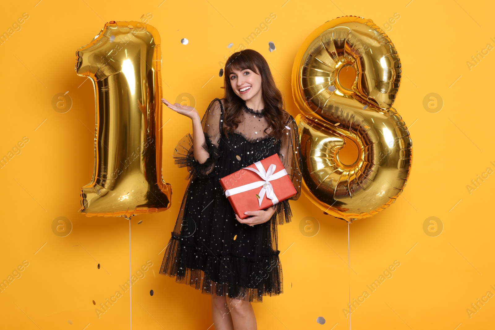 Photo of Coming of age party - 18th birthday. Young woman with number shaped balloons and gift box on yellow background