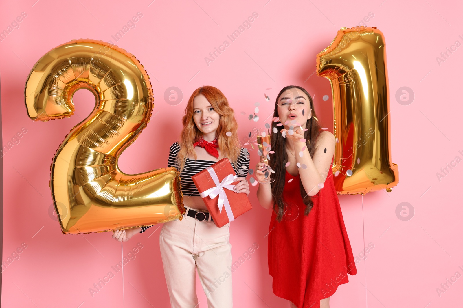 Photo of Coming of age party - 21st birthday. Young women celebrating with number shaped balloons and blowing confetti on pink background