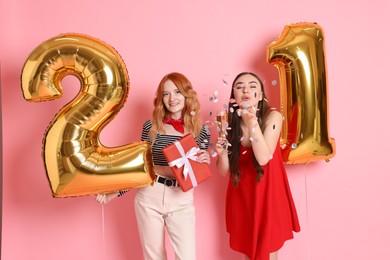 Photo of Coming of age party - 21st birthday. Young women celebrating with number shaped balloons and blowing confetti on pink background