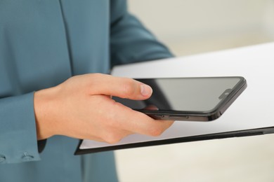 Photo of Businesswoman with clipboard using smartphone indoors, closeup. Modern technology