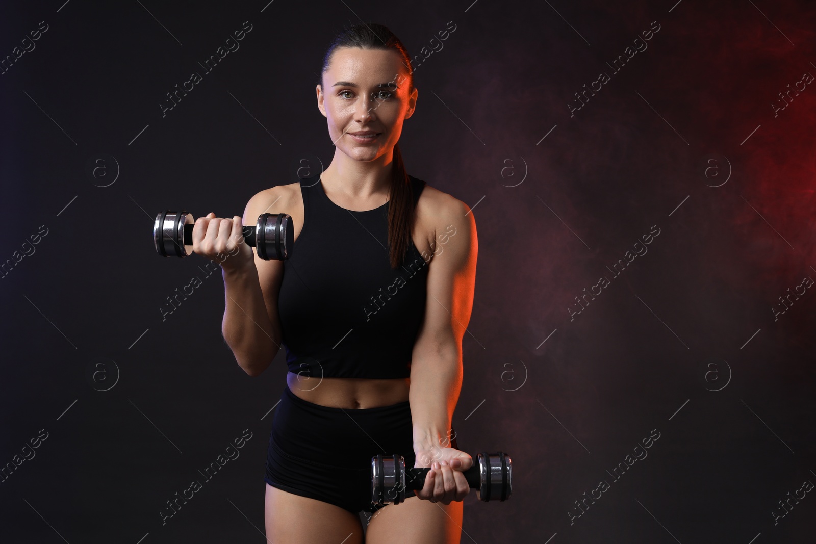 Photo of Woman exercising with dumbbells in smoke on dark background, space for text