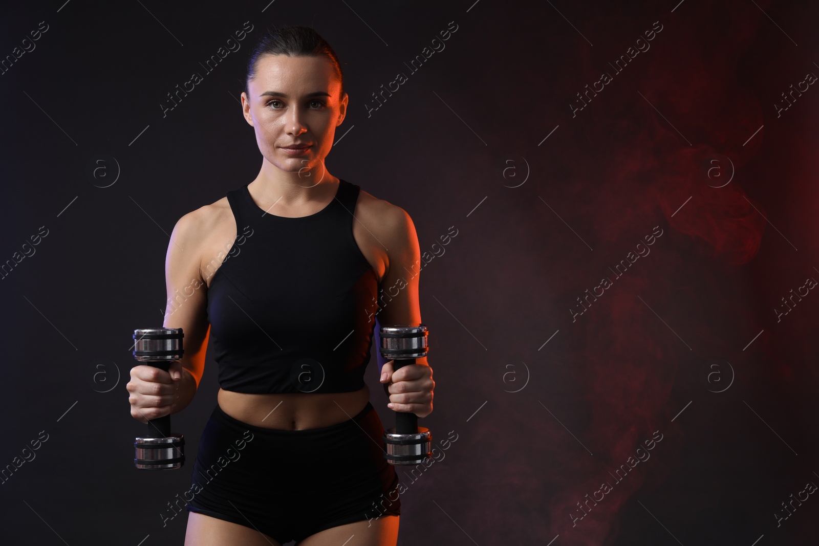 Photo of Woman exercising with dumbbells in smoke on dark background, space for text