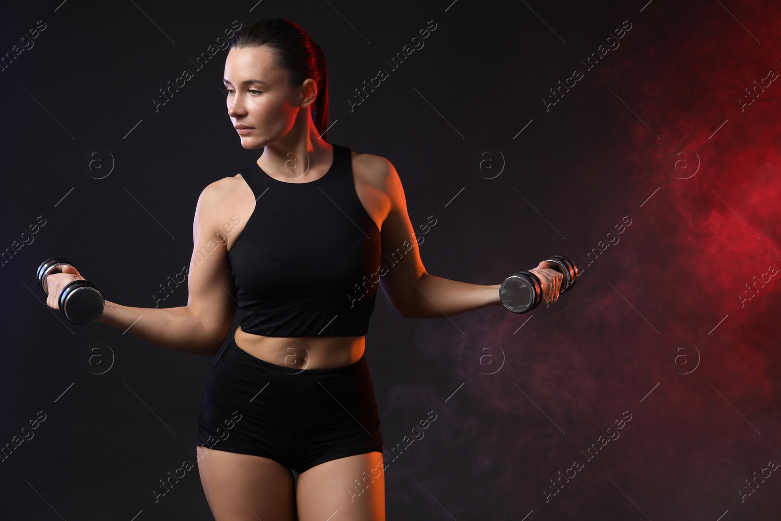 Photo of Woman exercising with dumbbells in smoke on dark background. Space for text