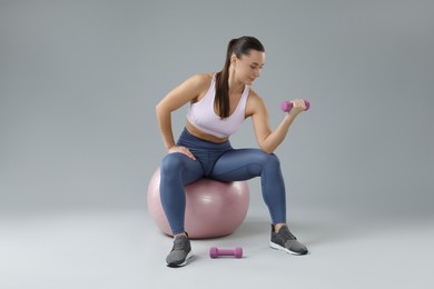 Woman exercising with dumbbells on fitball against light grey background