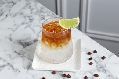 Refreshing espresso tonic drink with slice of lime and coffee beans on white marble table