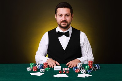 Photo of Professional croupier at gambling table with playing cards, casino chips and dice against dark yellow background