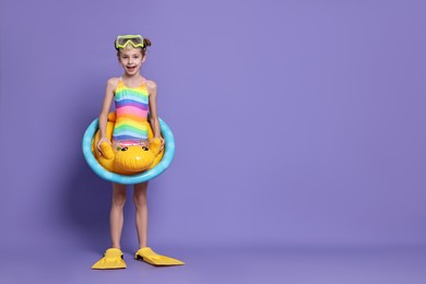 Photo of Happy girl in beachwear with diving mask, flippers and inflatable ring on purple background, space for text