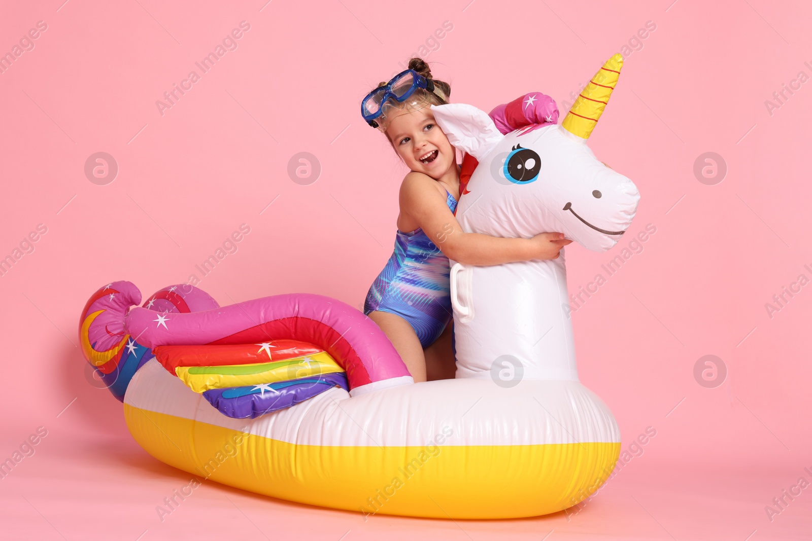 Photo of Cute little girl in swimsuit with unicorn pool float on light pink background