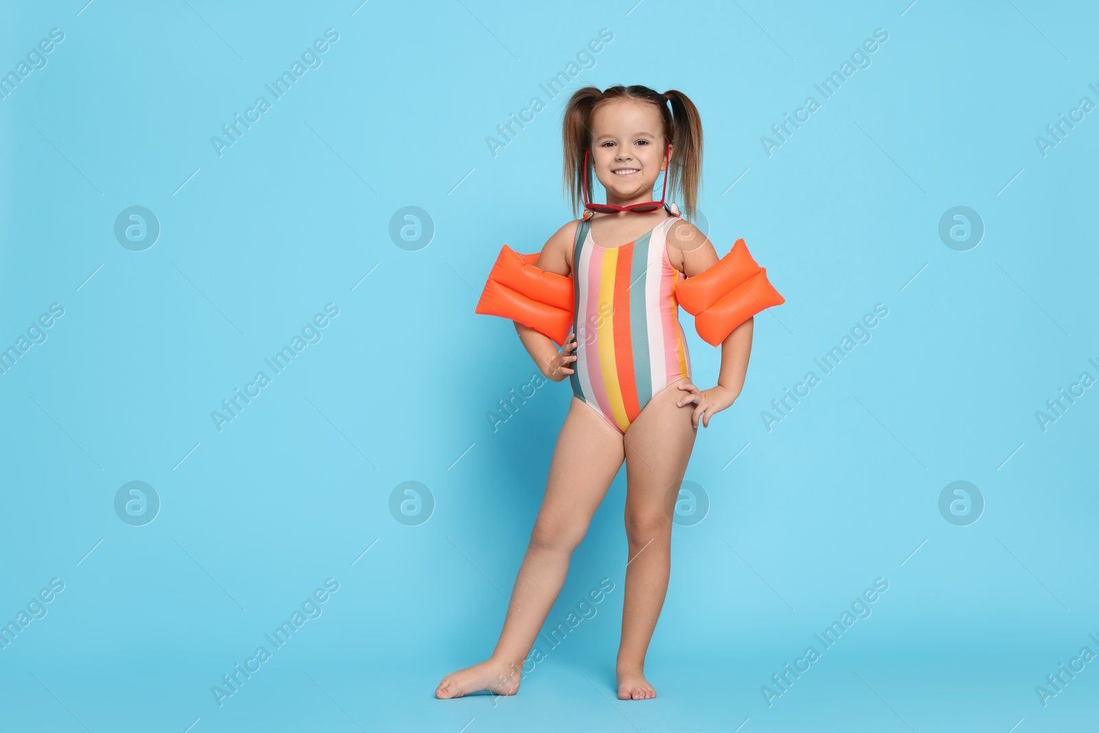 Photo of Cute little girl in swimsuit with swim armbands on light blue background