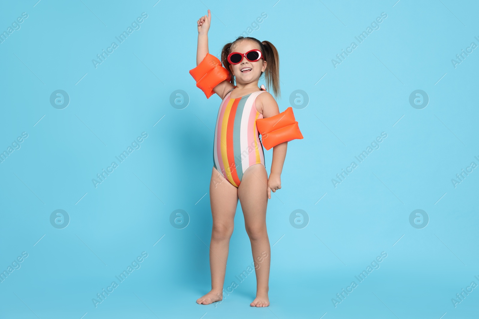 Photo of Cute little girl in swimsuit with swim armbands on light blue background