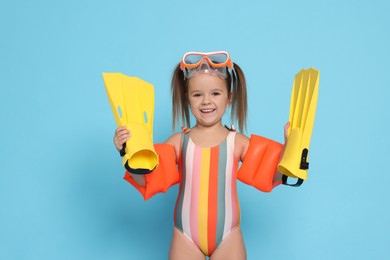 Photo of Cute little girl in swimsuit with swim armbands and fins on light blue background