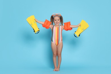 Photo of Cute little girl in swimsuit with swim armbands and fins on light blue background